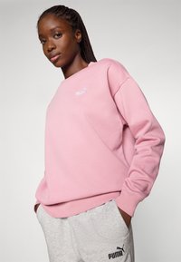 Felpa rosa con scollatura a girocollo e polsini a costine; pantaloni della tuta grigi visibili con un logo scuro. Tessuto morbido e strutturato.