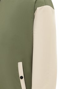 Olíva zöld dzseki bézs ujjaival, sima textúrával, letisztult vonalakkal és fekete gomb kiemeléssel a zsebén.