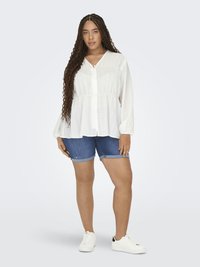 Kvinna med långt flätat hår som bär en vit blus med knappar, jeansshorts, vita sneakers och minimal smyckning, stående mot en enkel bakgrund.