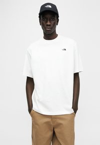 Λευκό βαμβακερό t-shirt με στρογγυλή λαιμόκοψη και κοντά μανίκια, με ένα μικρό μαύρο λογότυπο στο αριστερό στήθος. Συνδυάζεται με μπεζ παντελόνια και μαύρο καπέλο.