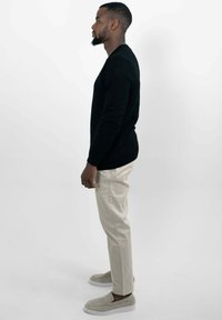 Zwarte gebreide trui, beige broek, lichtbeige instappers; van opzij weergegeven tegen een plain witte achtergrond.