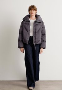 Grå puffjakke med høy krage, diagonal quiltmønster, frontglidelås og elastiske mansjetter. Kombinert med mørke denimbukser.