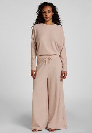 Vrouw met krullend haar, gekleed in een beige, langharige wafelgebreide top en bijpassende wijde broek met trekkoord in de taille, staand op blote voeten.