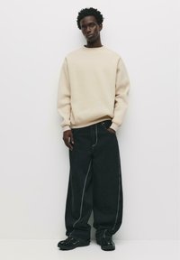 Sweatshirt beige a manica lunga con collo a giro, abbinato a jeans in denim neri oversize con cuciture bianche a contrasto. Sneakers nere.