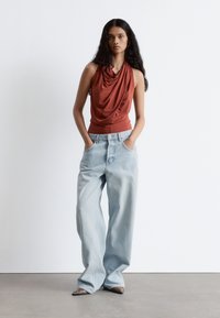 Rust-rotes ärmelloses Oberteil mit drapiertem Ausschnitt, kombiniert mit hellblauen weit geschnittenen Jeans. Silberne Akzente an den Jeans und strukturierte Schuhe.