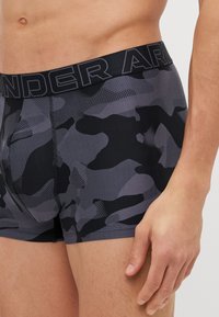 Boxers para hombres en un patrón de camuflaje negro y gris, con una cinturilla negra sólida que lleva el logo 'UNDER ARMOUR', material elástico suave.