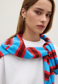Multiculturele sjaal met gedurfde horizontale strepen in blauw, rood en bruin, gemaakt van zachte stof, om de nek gedrapeerd.