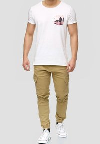 Weißes T-Shirt mit kurzen Ärmeln und einem pinken Palmen-Design auf der Brusttasche, kombiniert mit beigen Cargo-Hosen und weißen Sneakers.