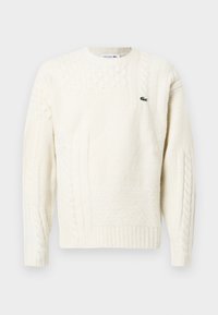 Krémový pletený sveter s variáciami textúry, obsahujúci káblový dizajn, rebrované manžety a malé zelené logo Lacoste na hrudi.