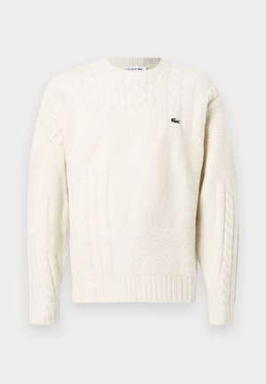 Krém színű kötött pulóver, textúra változásokkal, kábelmintával, bordás mandzsettákkal és egy kis zöld Lacoste logóval a mellrészén.