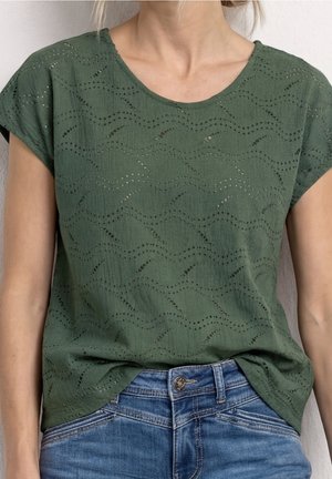 Femme portant un chemisier vert à manches courtes avec des motifs découpés en forme de vagues, rentré dans un jean en denim bleu.