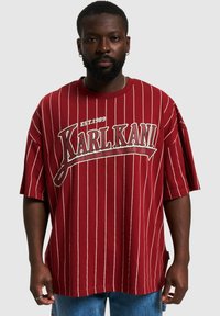 T-shirt oversize rossa con strisce verticali bianche, con logo "KARL KANI EST.1989" in testo in grassetto. Tessuto morbido, vestibilità rilassata.