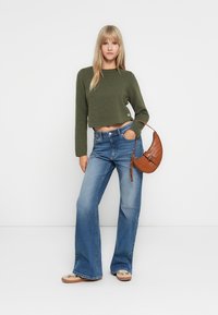 Maglione verde corto con maniche lunghe, jeans blu a vita alta a gamba larga e borsa in pelle marrone, indossati con scarpe beige casual.