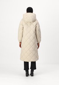 Lange, lichtbeige pufferjas met een doorgestikt ruitpatroon, een oversized capuchon en elastische manchetten. Draagt over zwarte textuurbroek en chunky hakken.