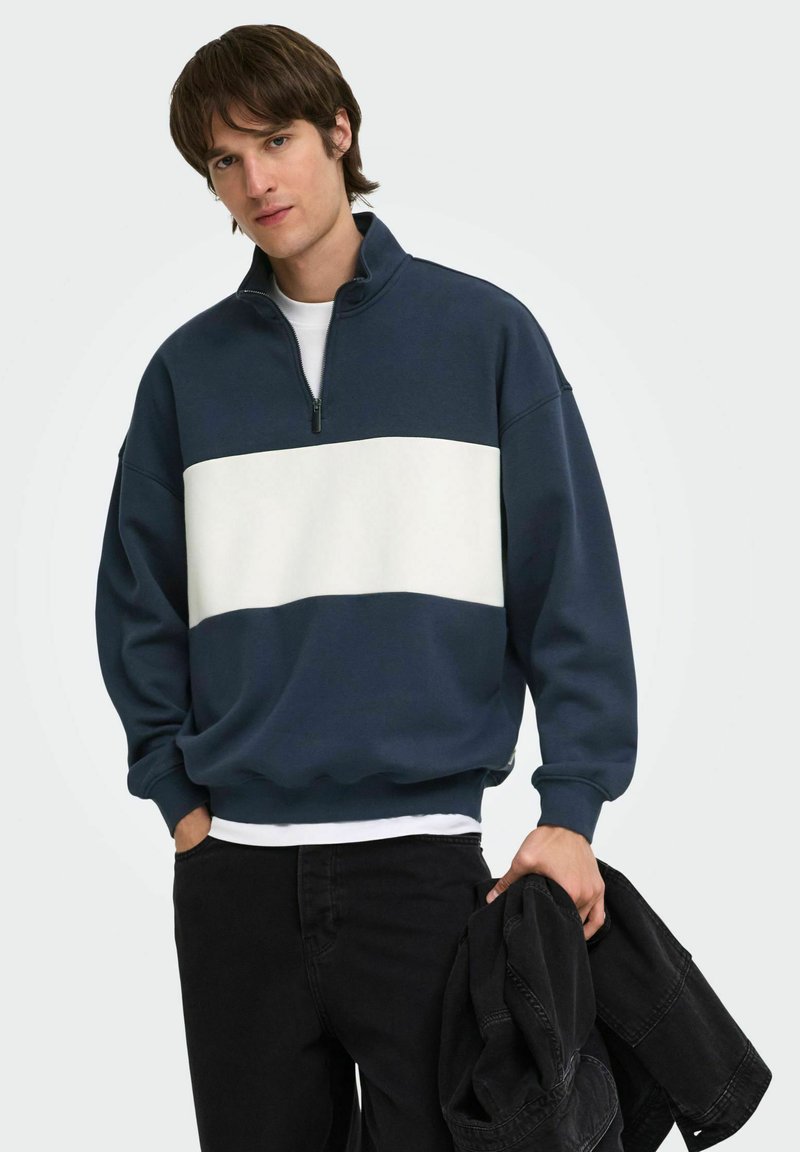 Jeune homme portant un sweat-shirt marine et blanc à fermeture éclair quart, pantalon noir, tenant une veste noire dans la main gauche, debout devant un fond uni.