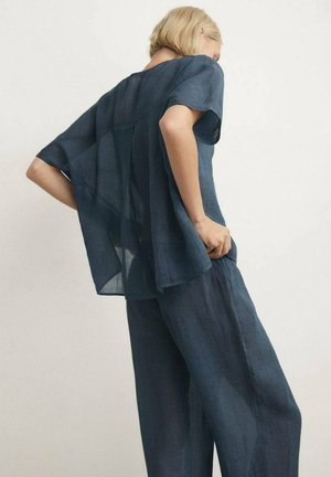 Mujer con cabello corto y rubio, vestida con un top suelto y translúcido azul marino de manga corta y pantalones anchos a juego, de pie con las manos en las caderas.