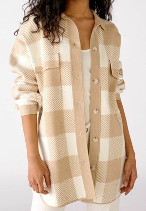 Frau trägt einen beige-weiß karierten, geknöpften Cardigan mit hochgekrempelten Ärmeln über einem weißen Top und einer weißen Hose.