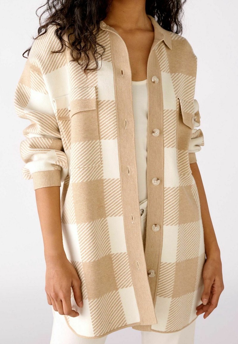 Femme portant un cardigan boutonné à carreaux beige et blanc avec les manches retroussées sur un haut blanc et un pantalon.
