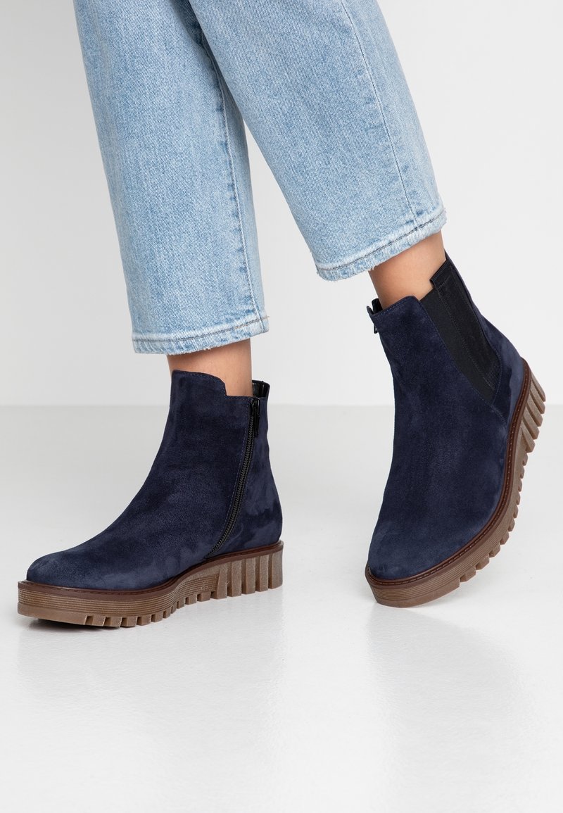 Chevilles avec des jeans bleu clair courts portant des bottines en daim bleu marine avec des semelles épaisses et côtelées marron et des fermetures éclair latérales, debout sur un sol blanc.