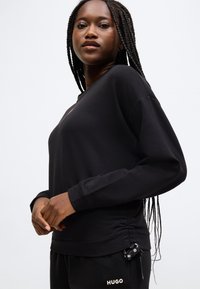 Musta collegepaita, joka on valmistettu pehmeästä kankaasta. Siinä on pitkät hihat, pyöreä kaula-aukko ja sivustassa kiristysnyörit, jotka mahdollistavat säädettävän istuvuuden.