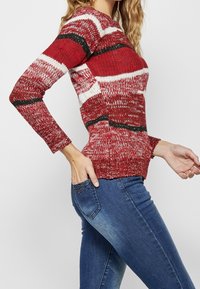 Roter gestrickter Pullover mit weißen und schwarzen gestreiften Mustern; strukturierter Stoff; taillierte Passform; getragen mit blauen Jeans mit verzierten Taschen.