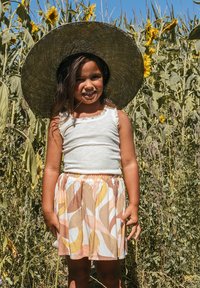 Kind met een wit ribbel topje en een kleurrijke rok met draaien in geel, roze en bruin; grote strohoed; zonnebloemen op de achtergrond.