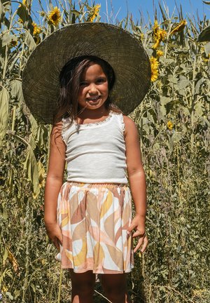 Kind met een wit ribbel topje en een kleurrijke rok met draaien in geel, roze en bruin; grote strohoed; zonnebloemen op de achtergrond.