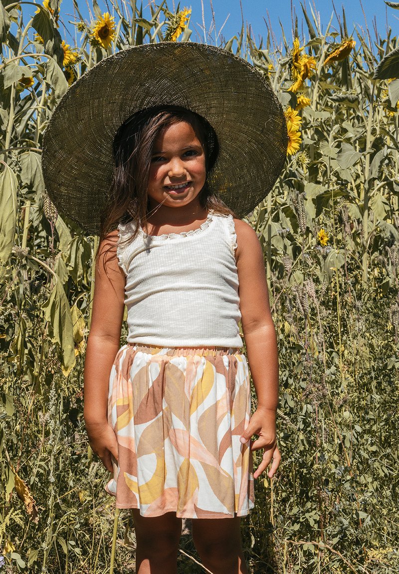 Kind met een wit ribbel topje en een kleurrijke rok met draaien in geel, roze en bruin; grote strohoed; zonnebloemen op de achtergrond.