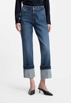 Person iført mørkeblå brede cowboybukser med opkrogede bukseben, sort langærmet top og sorte kitten heel mule, stående på hvid baggrund.