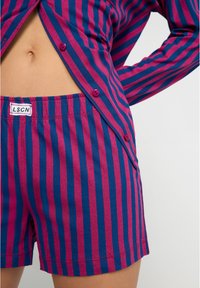 Calções às riscas em algodão azul e magenta com cintura elástica e etiqueta da marca. Apresenta botões numa camisa correspondente nas proximidades.