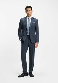 Donkerblauw op maat gemaakt pak met inkeping revers, een wit overhemd en een lichtblauwe stropdas. Gepolijste zwarte schoenen maken de look compleet.