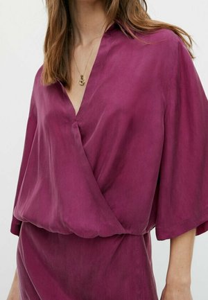 Woman wearing a loose-fitting, deep purple V-neck blouse with three-quarter sleeves and a gold pendant necklace.
