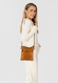 Borsa a tracolla in pelle color nocciola con tasca frontale con zip, tracolla regolabile e superficie liscia, tenuta da una persona in abbigliamento casual.