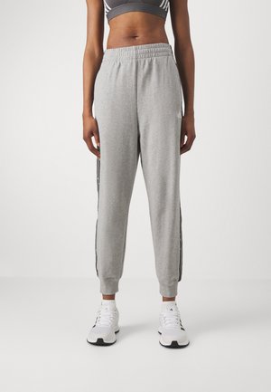 Woman wearing gray jogger pants with black side stripes, white sneakers, and a black-gray sports bra standing on a white floor against a white background.