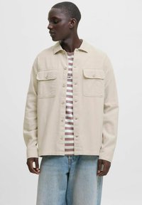 Beige gombos ing, két zsebbel a mellrészen, csíkos póló fölött viselve. A modellen laza kék farmernadrág van, ami egy kényelmes fazont mutat.