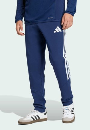 Mężczyzna noszący granatowe spodnie dresowe Adidas z białymi paskami i logo, w połączeniu z białymi trampkami z czarnymi paskami.