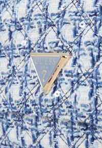 Tessuto testurizzato blu e bianco con paillettes presenta un distintivo triangolare con bordo dorato del marchio Guess U.S.A. al centro.