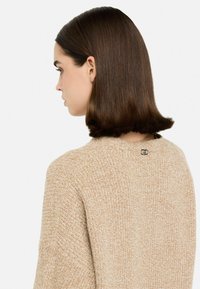 Beidžs, teksturēts džemperis ar apaļu kaklu un mazu sudraba logotipu aizmugurē. Mīksts audums ar mājīgu izskatu.