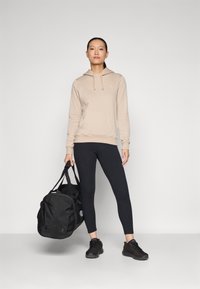 Sweat à capuche beige clair avec poche kangourou, associé à des leggings noirs. Le modèle tient un sac de sport noir et porte des baskets noires.