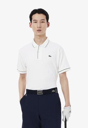 Polo bianca con colletto e bordo maniche verdi, con un piccolo logo coccodrillo ricamato. Indossata con pantaloni blu navy e una cintura coordinata.