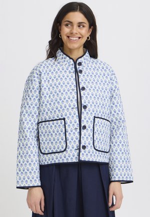 Femme souriante, portant une veste matelassée blanche avec un motif floral bleu, des bordures noires, de grandes poches avant, et une jupe bleu foncé.