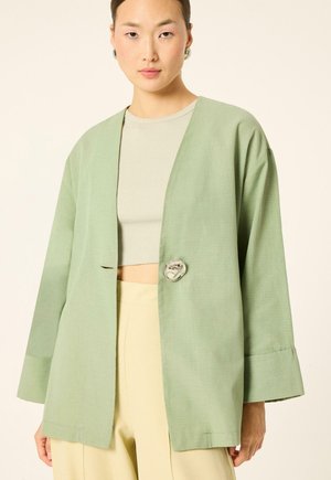 Femme portant un blazer oversize vert clair avec un grand bouton métallique, un crop top beige et un pantalon jaune clair sur un fond uni.