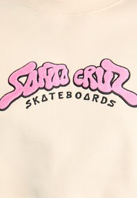 Felpa beige chiara con testo ricamato in rosa bold, "Santa Cruz," con superficie testurizzata. Sotto, "SKATEBOARDS" in nero.