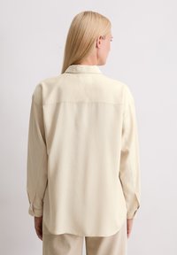 Helles beige Langarmhemd mit Kragen, das eine glatte Textur und einen leicht geschwungenen Saum aufweist. Mit Knopfbündchen und einem schlichten Design.