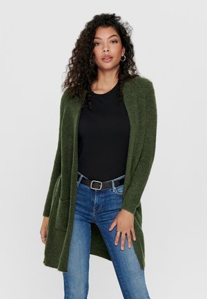 Cardigan verde in maglia con maniche lunghe, apertura frontale e due tasche laterali, indossato sopra una maglietta nera e jeans blu. Tessuto morbido, vestibilità comoda.