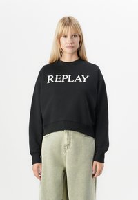 Blonde vrouw die een zwart sweatshirt met de tekst "REPLAY" en lichtgrijze high-waisted losse jeans draagt, tegen een plain witte achtergrond.