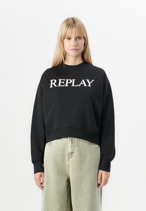Blonde vrouw die een zwart sweatshirt met de tekst "REPLAY" en lichtgrijze high-waisted losse jeans draagt, tegen een plain witte achtergrond.