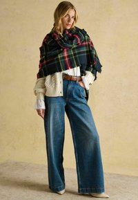 Villane ruuduline sall tumesinisel, punasel ja rohelisel heledale palmikumustriga kampsunile visatud. Laialäärmelised sinised teksad ja beežid kingad täiendavad välimust.