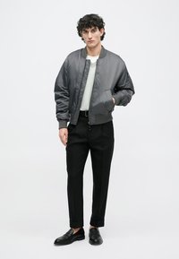 Young man with curly hair wearing a gray bomber jacket, light sweater, black trousers with cuffs, and black loafers, standing against white background.