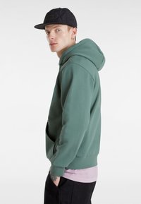 Felpa verde con cappuccio in un tessuto morbido, con tasca frontale e polsini a coste. Indossata sopra una maglietta rosa chiaro e abbinata a pantaloni neri.